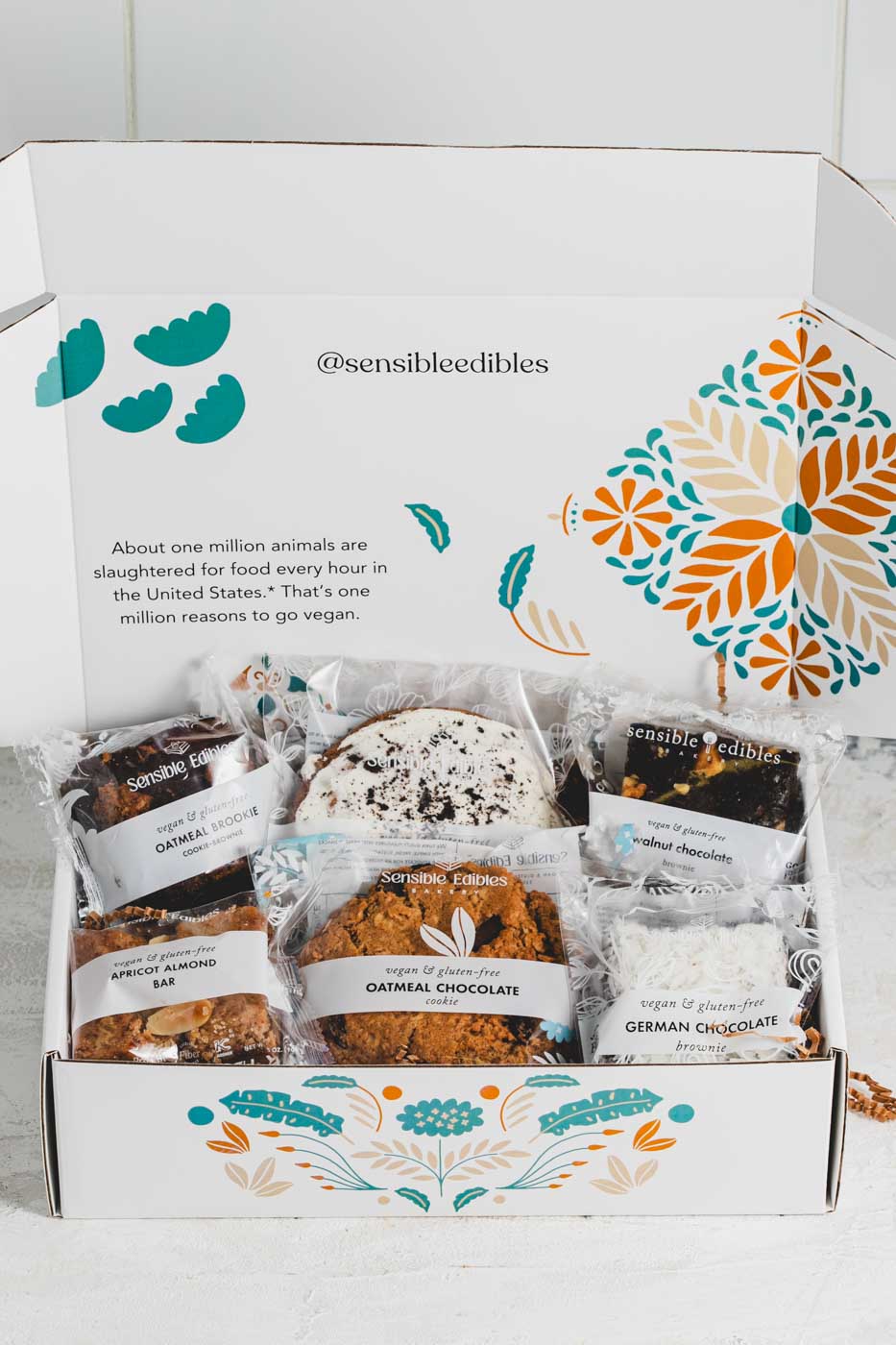 An open white gift box with colorful floral patters  displaying an assortment of packaged food items, including an oatmeal brookie, an apricot almont bar, a walnut chocolate brownie, a german chocolate brownie, an oatmeal chocolate cookie, and a cookie crumble cookie