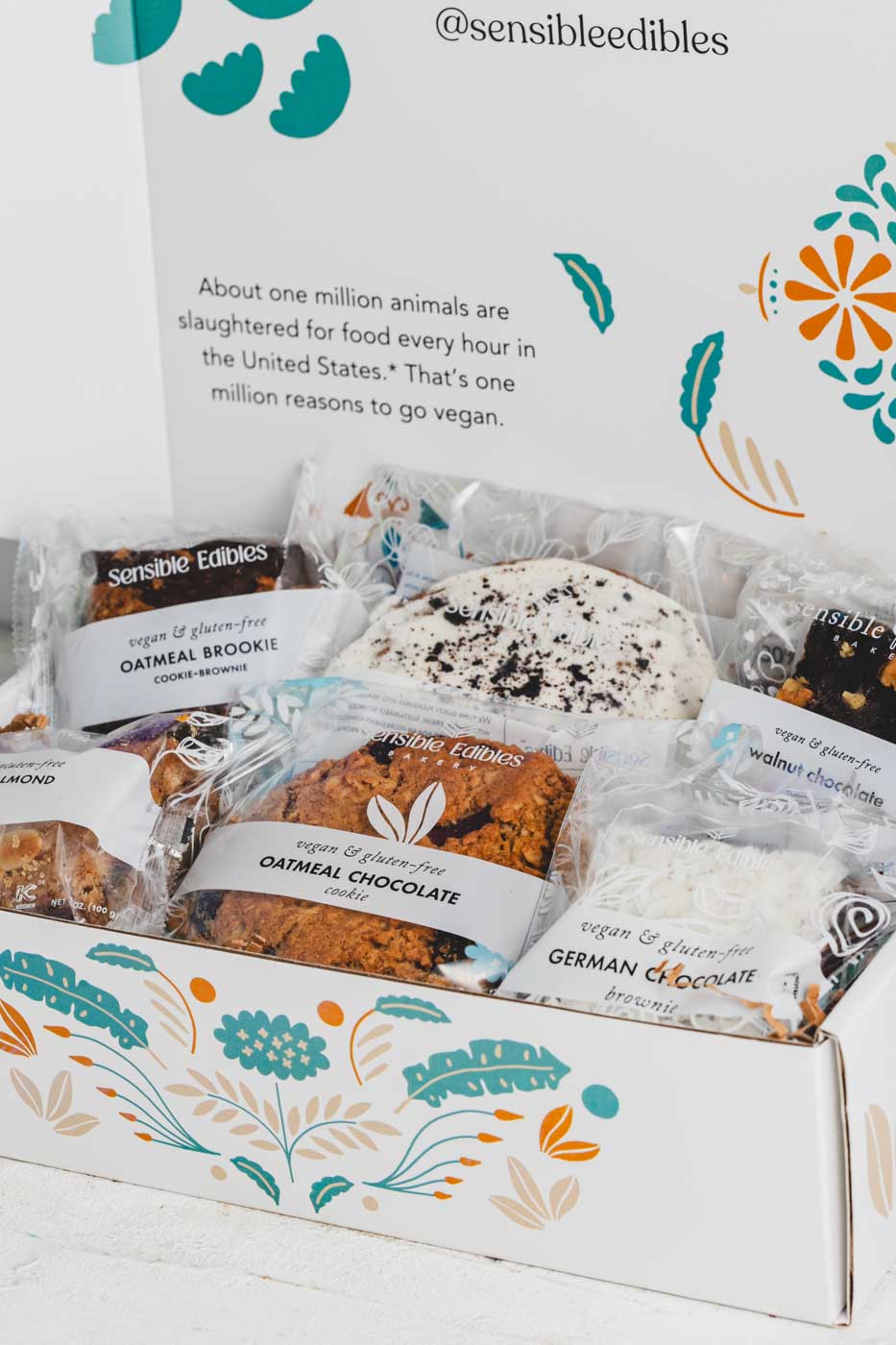 An open white gift box with colorful floral patters  displaying an assortment of packaged food items, including an oatmeal brookie, an apricot almont bar, a walnut chocolate brownie, a german chocolate brownie, an oatmeal chocolate cookie, and a cookie crumble cookie