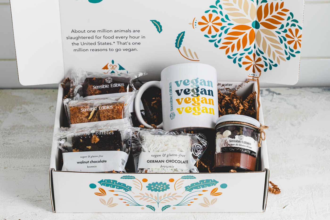 An open gift box containin various bars and brownies as well as a jar of hot chocolate mix and a mug with vegan written on it. The box is white with a floral pattern.