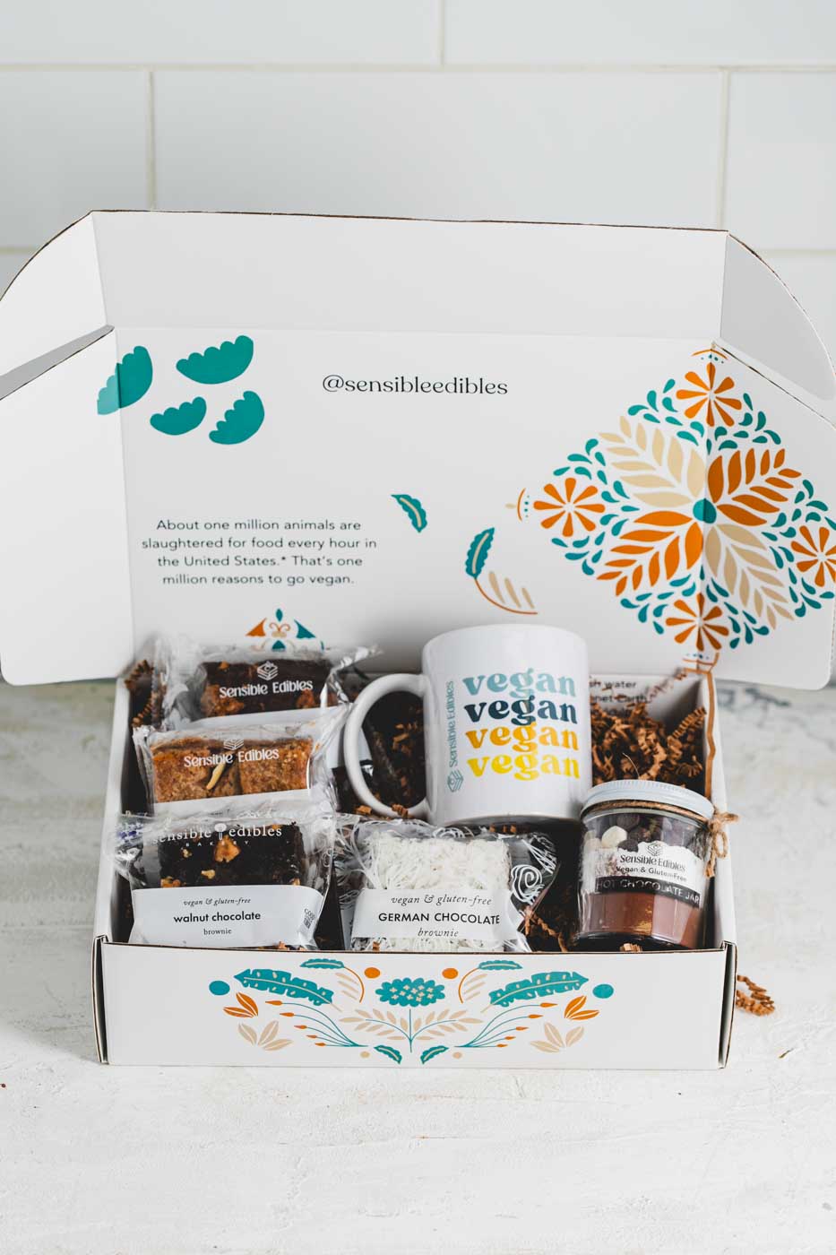 An open gift box containin various bars and brownies as well as a jar of hot chocolate mix and a mug with vegan written on it. The box is white with a floral pattern.
