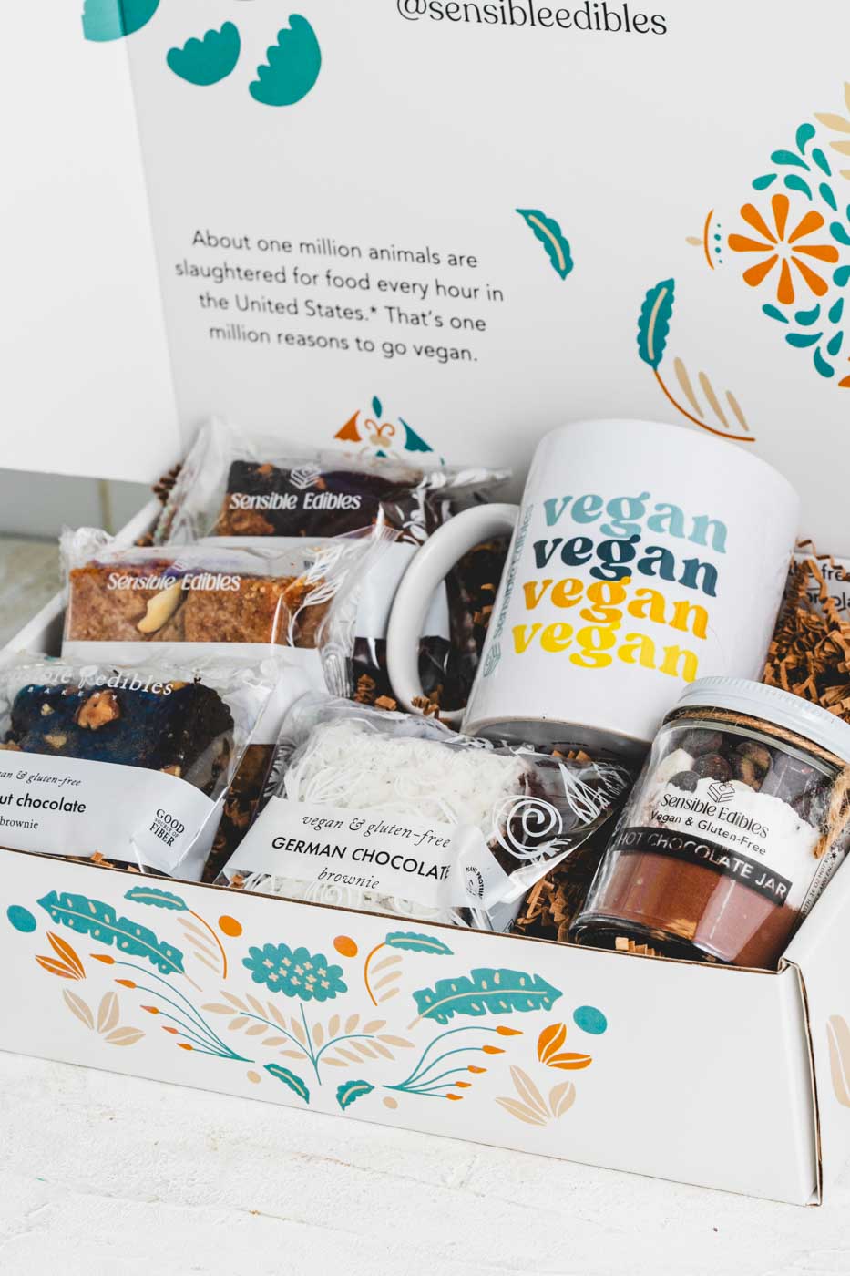 An open gift box containin various bars and brownies as well as a jar of hot chocolate mix and a mug with vegan written on it. The box is white with a floral pattern.