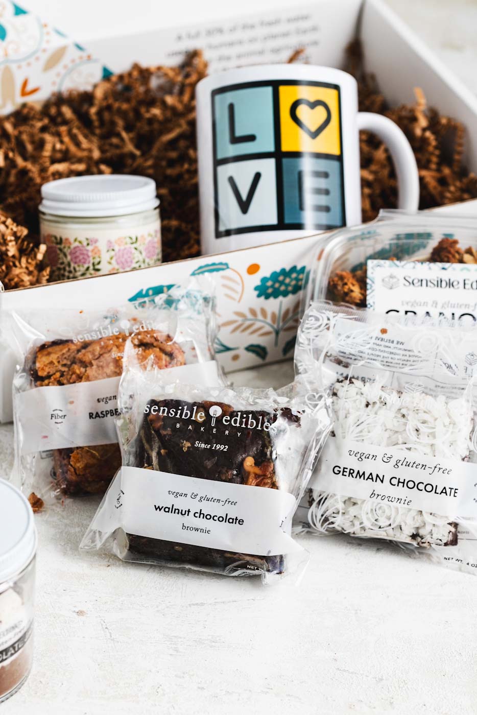 Gift box with contents partially taken out in front of it: Jar of hot chocolate mix, walnut chocolate brownie, sensible edibles mug, candle, granola, and german chocolate brownie.