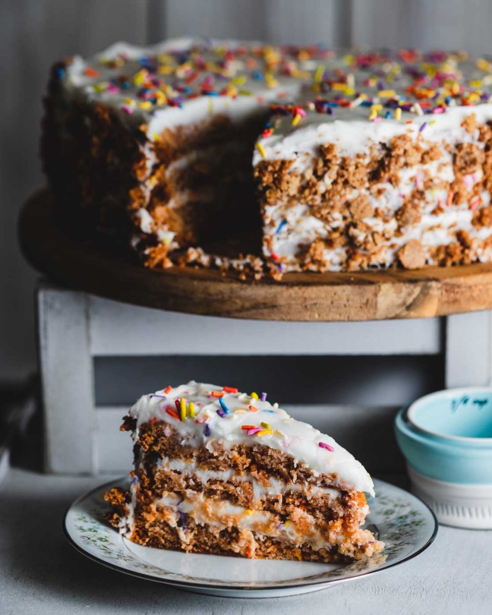 Slice of vanilla birthday cake with white frosting and colorful sprinkles on a floral plate, with the whole cake on a wooden stand in the background.
