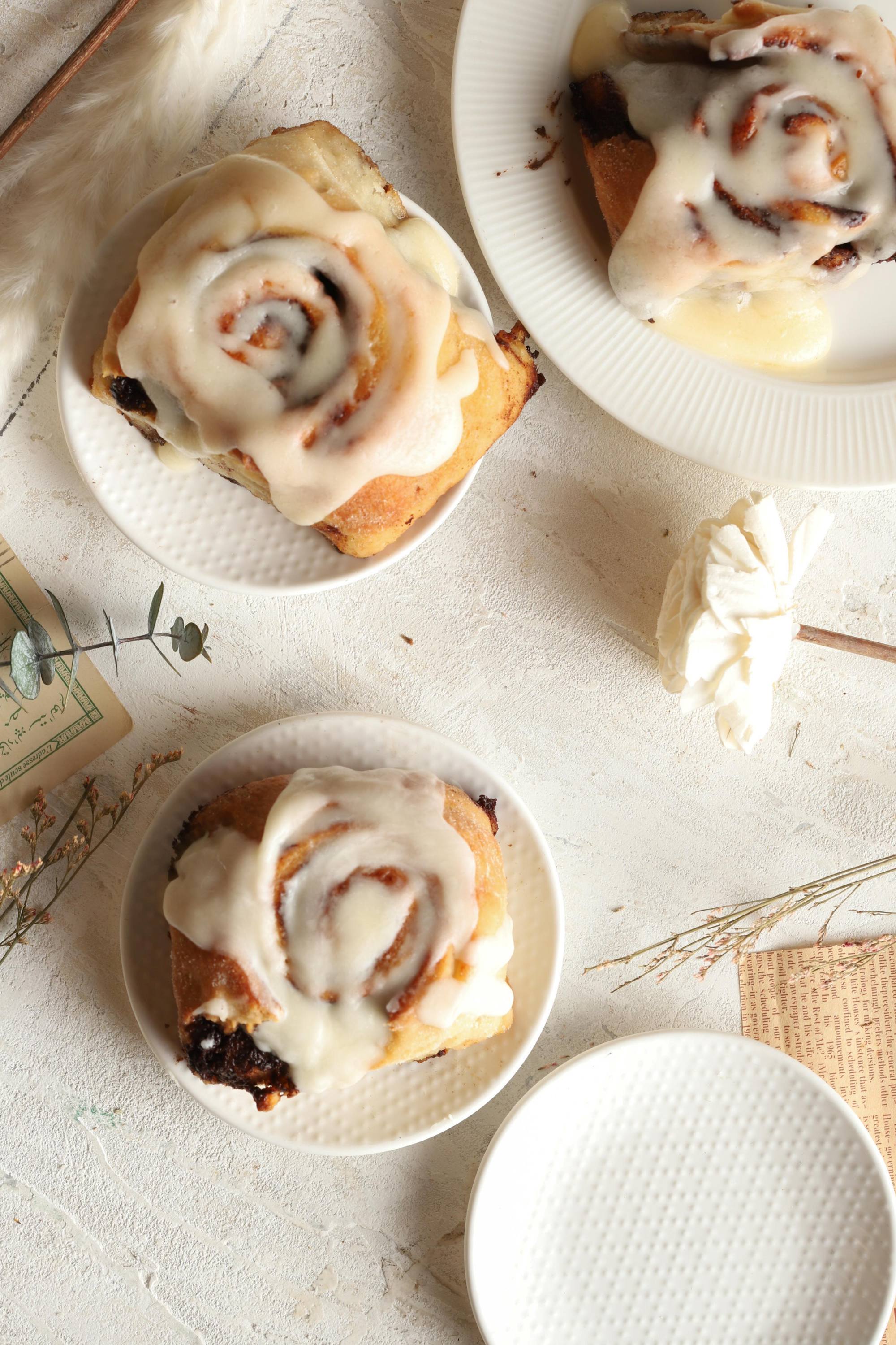 Cinnamon rolls with white icing on small white plates on a light surface.