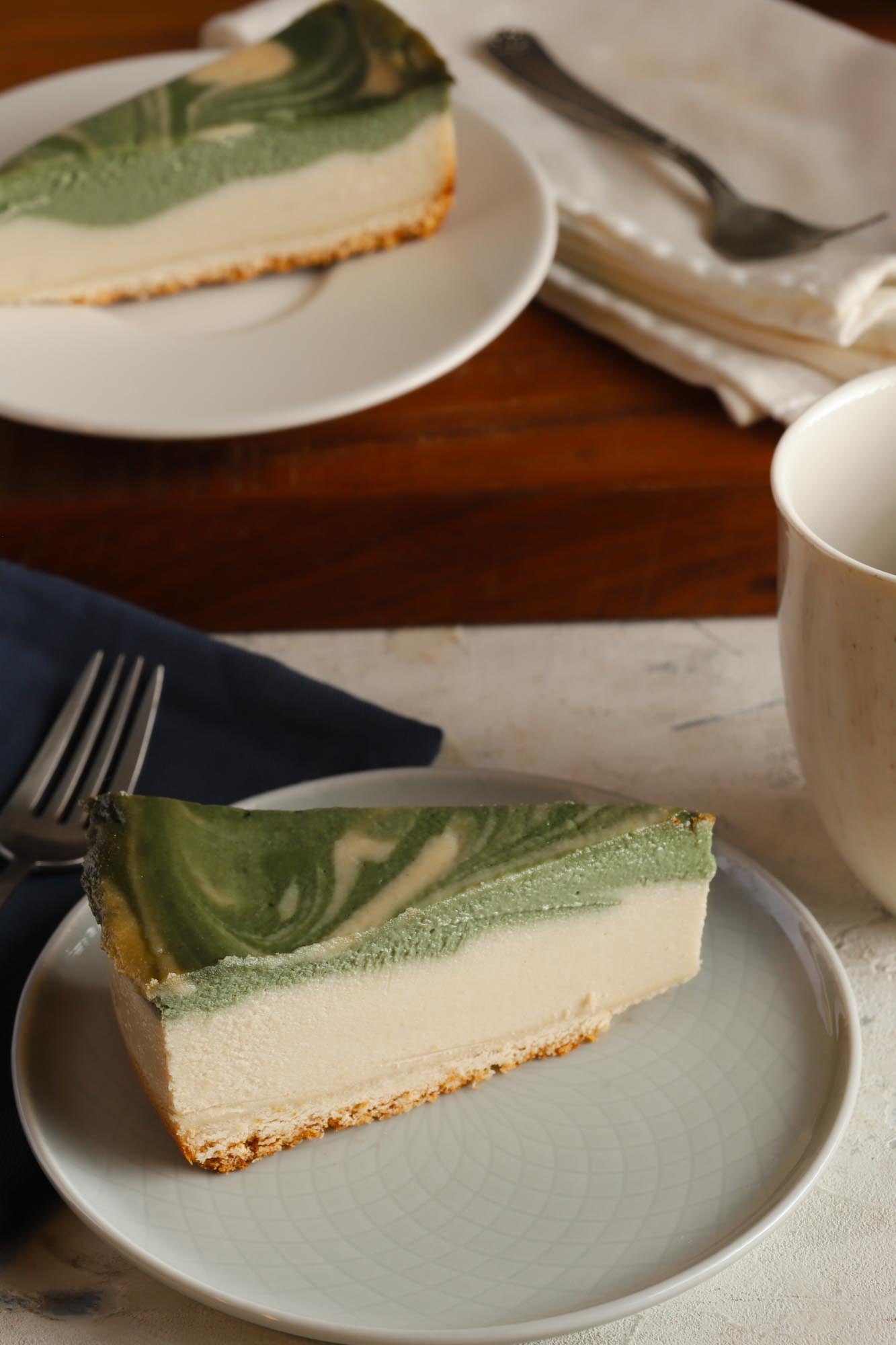 A slice of matcha cheesecake on a plate with a fork and mug and more slices in the background