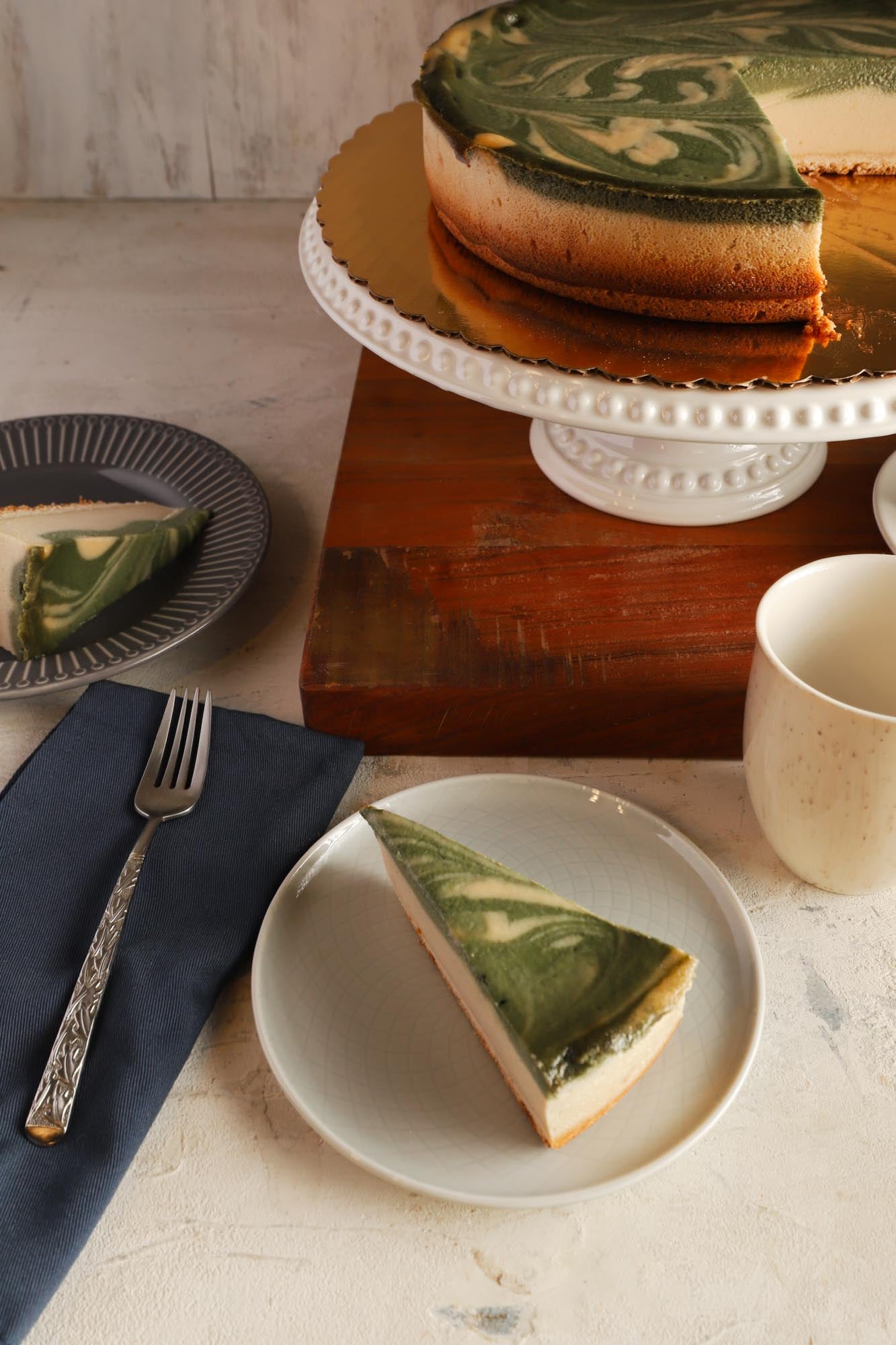 A slice on matcha cheesecake on a plate with more slices behind it and the rest of the cheesecake on a cake stand