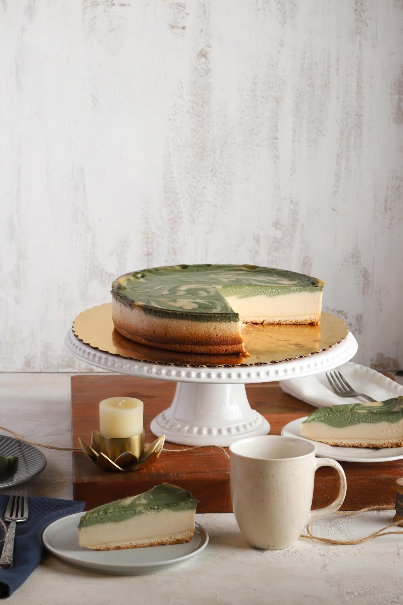 A vegan gluten-free matcha cheesecake on a cake stand with slices on plates and table settings around it