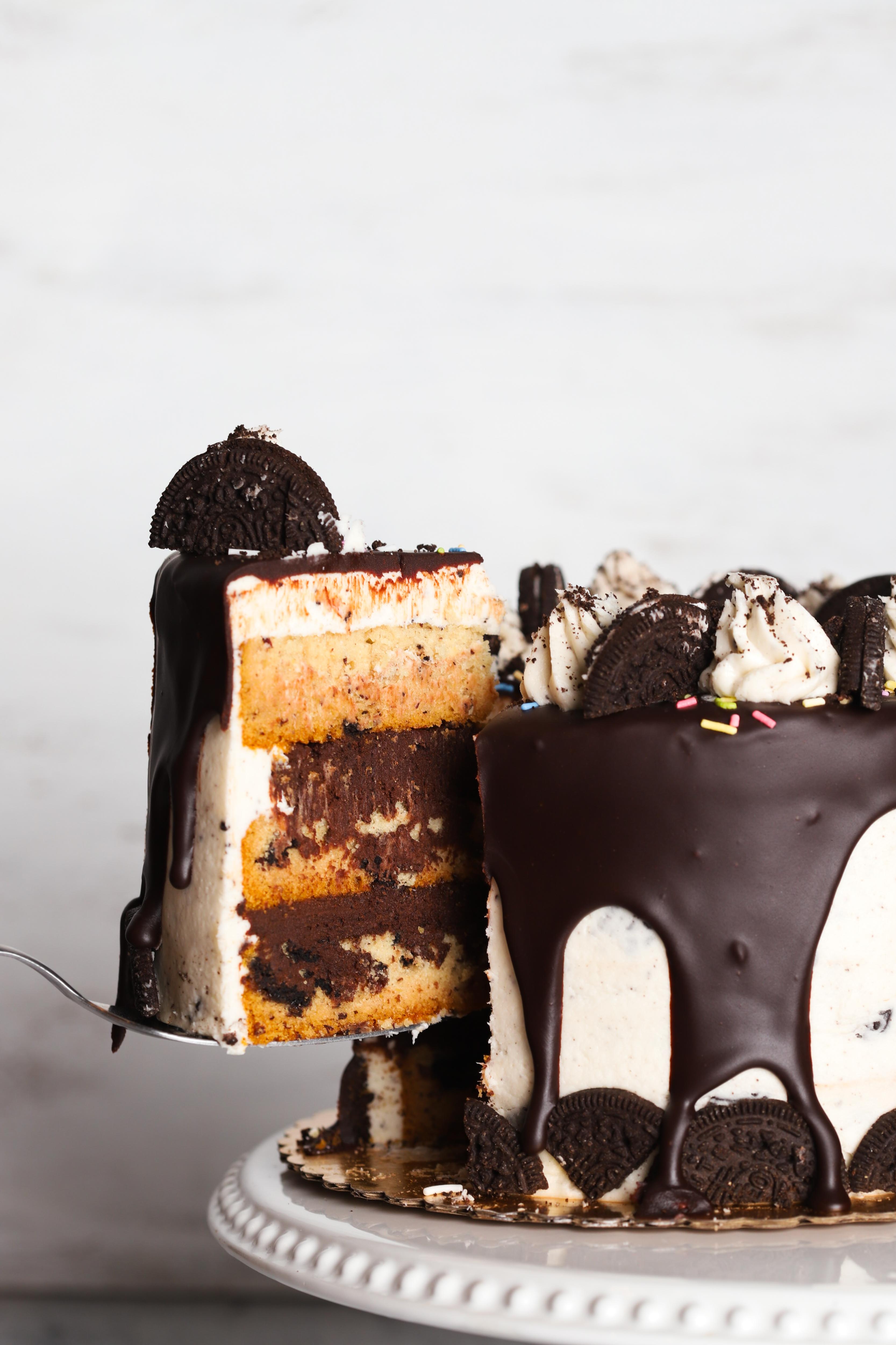 Cookies and cream cake with vanilla and chocolate frosting decorated with oreo cookies, sprinkles, and cookie crumbs with a slice being lifted out