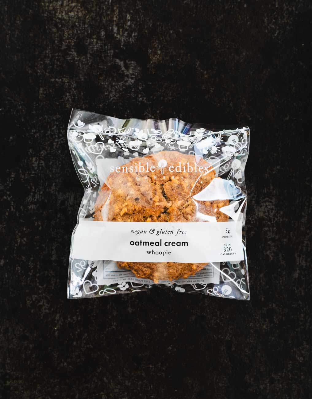 Packaged oatmeal cream whoopie from Sensible Edibles Bakery, labeled as vegan and gluten-free, on a dark background.