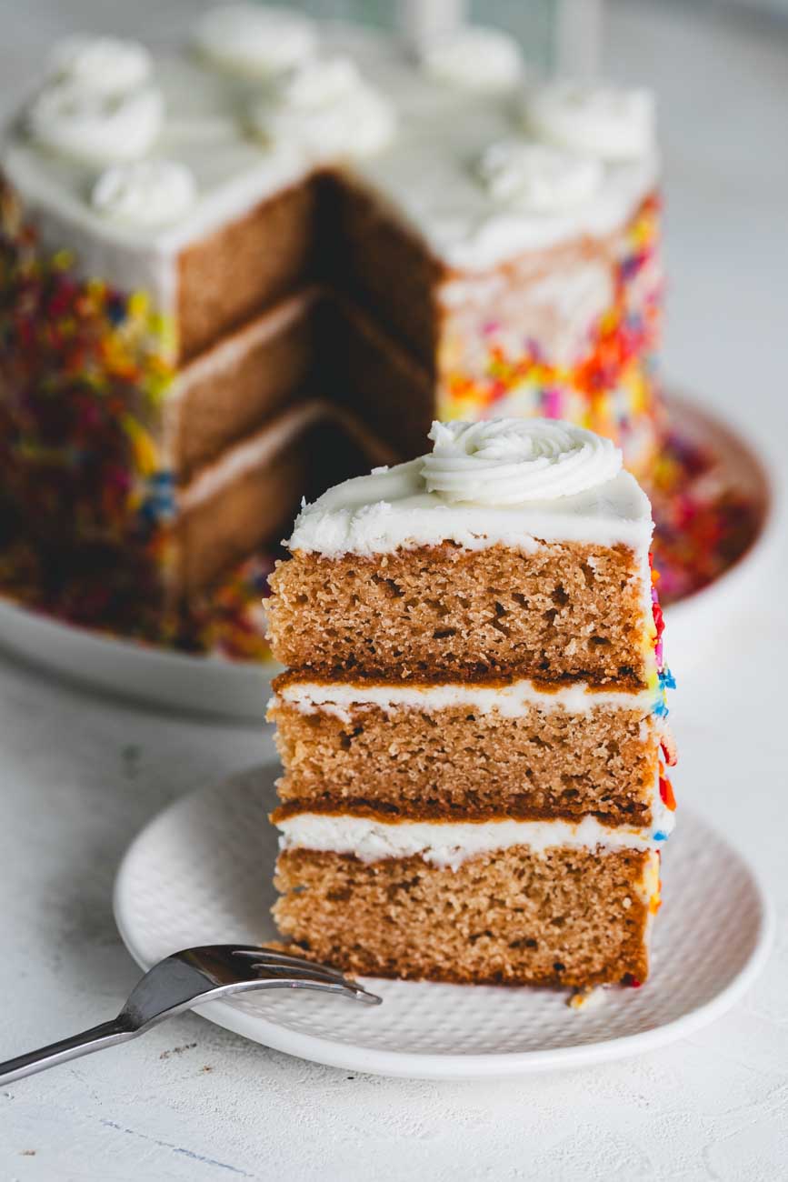 6-inch Vanilla birthday cake with white frosting and rainbow sprinkles. A slice is taken out on a plate with a fork.