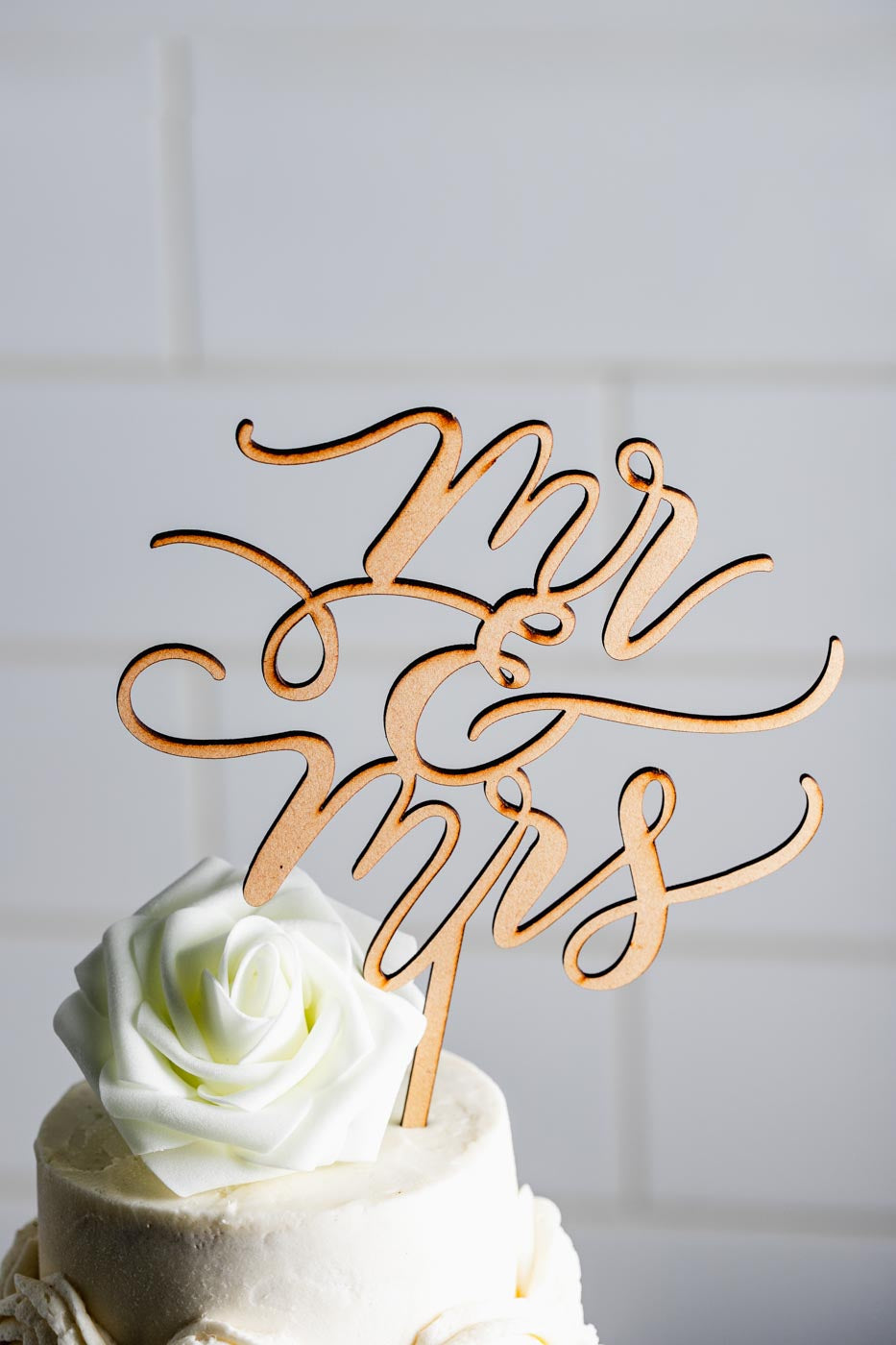 Close up of the top of a white tiered wedding cake with a white flower on top and a gold cake topper that says "mr & mrs"