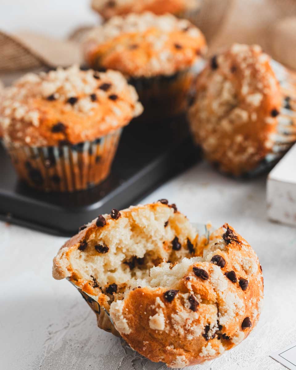 Chocolate chip muffins with a crumb topping, one muffin broken open to show the fluffy interior, arranged on a light surface with additional muffins in the background.