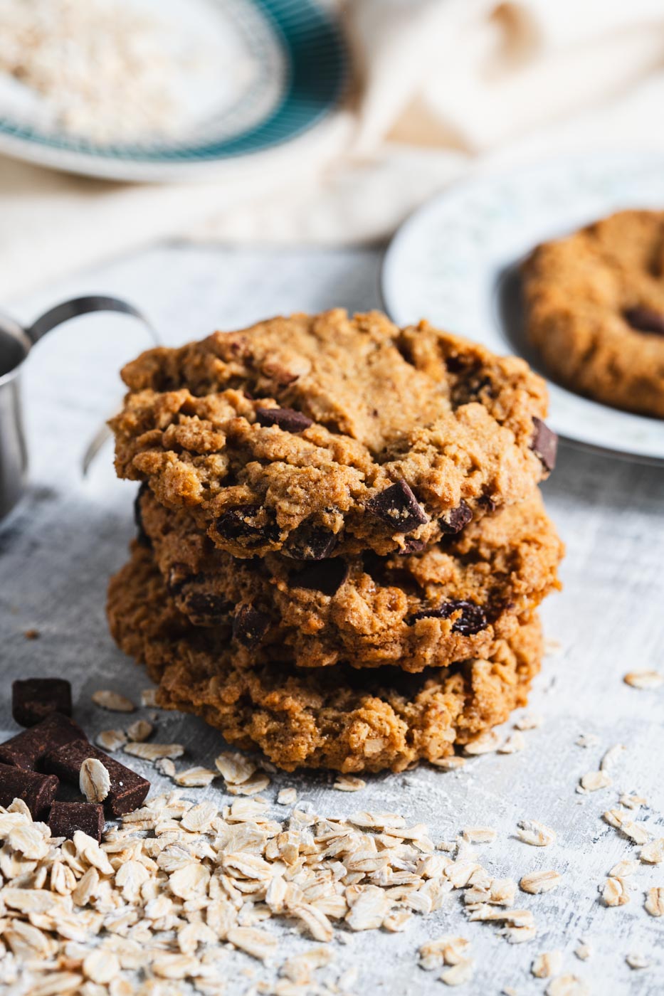 Stack of oatmeal chocolate chunk cookies with visible oats and chocolate pieces, on a light surface with a plate of cookies in the background.