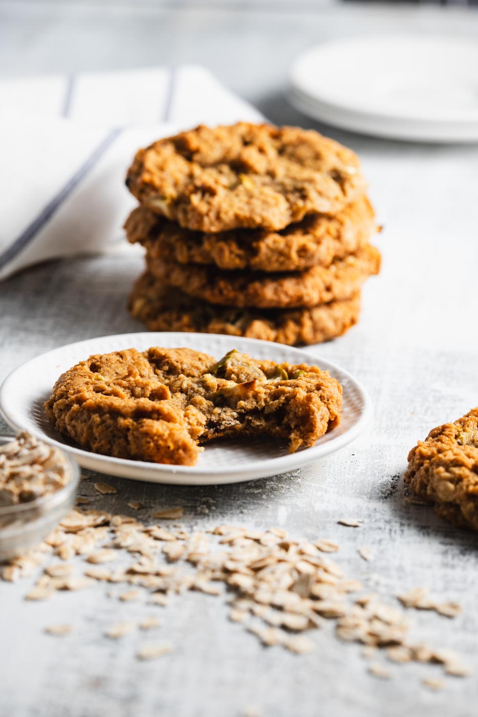 Stack of apple oatmeal cookies with one cookie on a plate with a bite taken out of it, showcasing the texture and apples inside. The cookies are on a light background with scattered oats around.