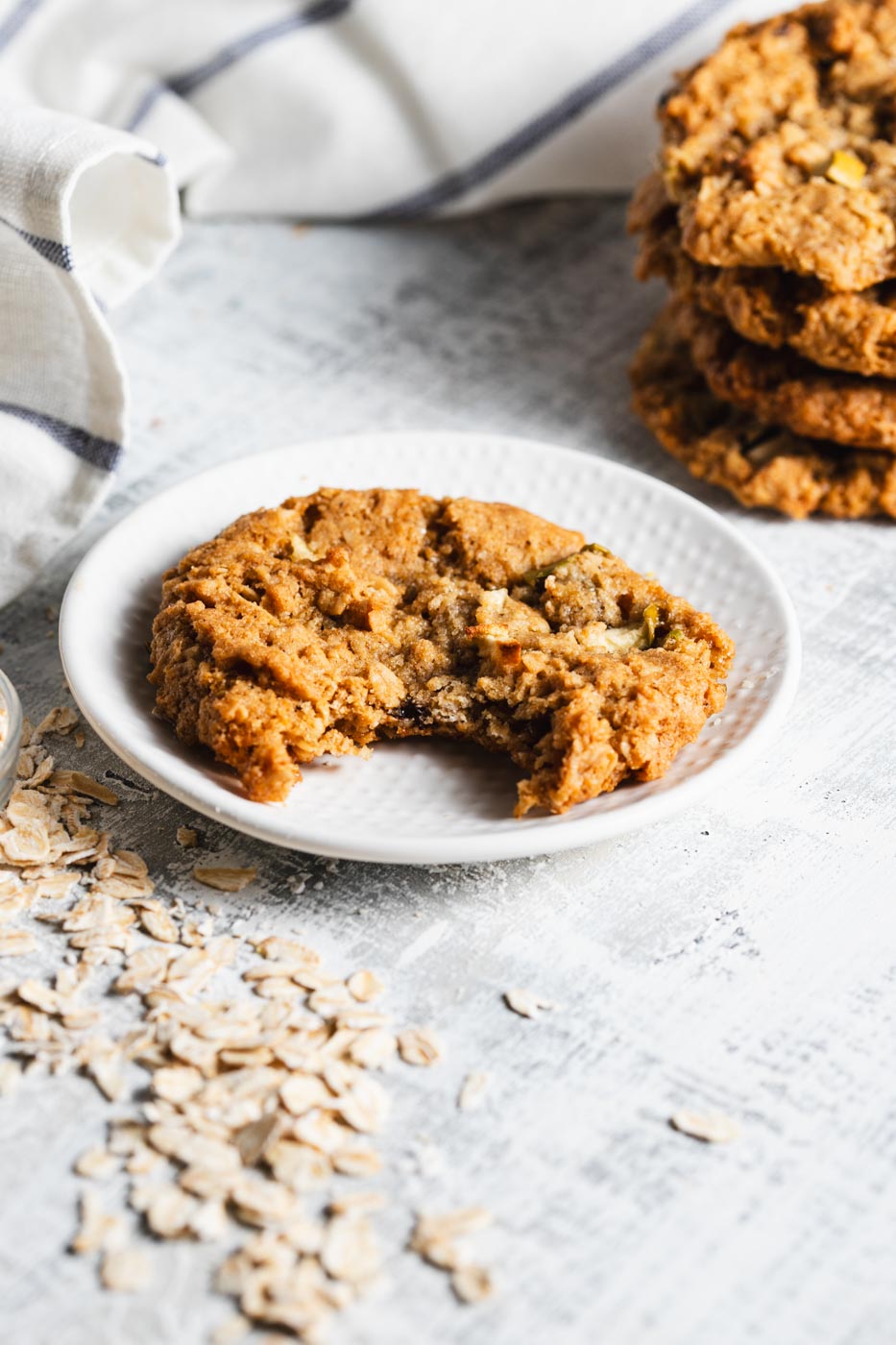 An apple oatmeal cookie on a white plate with a bite taken out of it. There is a stack of more cookies behing it and oats piled around.