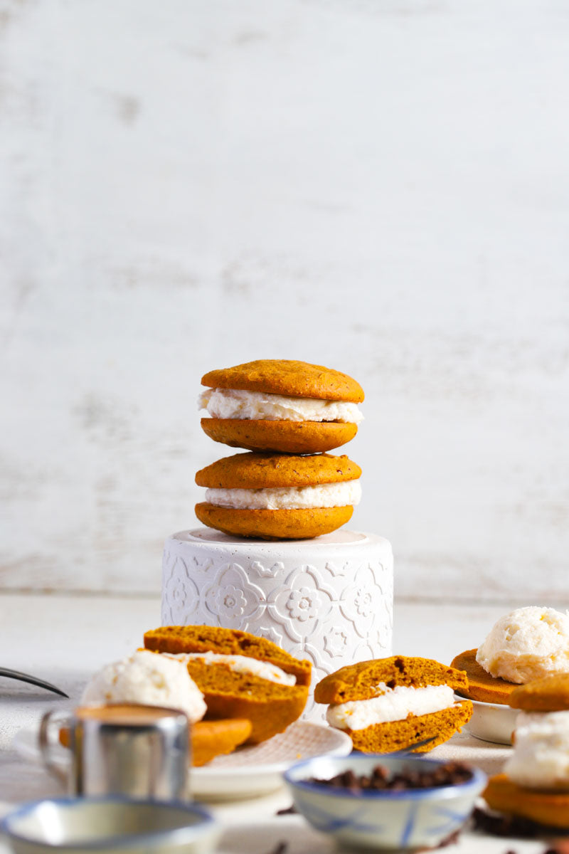 Pumpkin spice whoopies on and around  white plates with spices and coffee