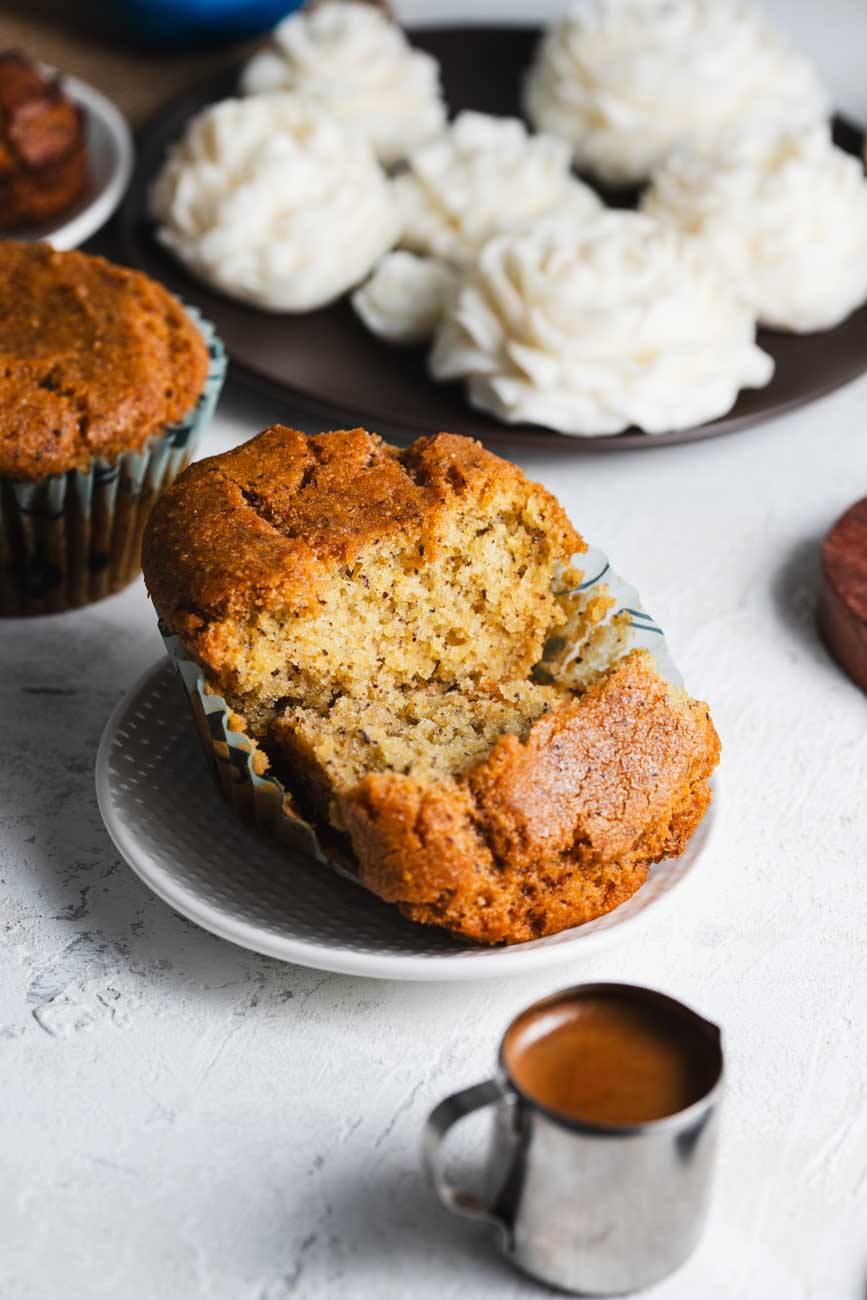 A single muffin broken open to reveal a moist, textured inside, placed on a white plate with other muffins and white flowers in the background.