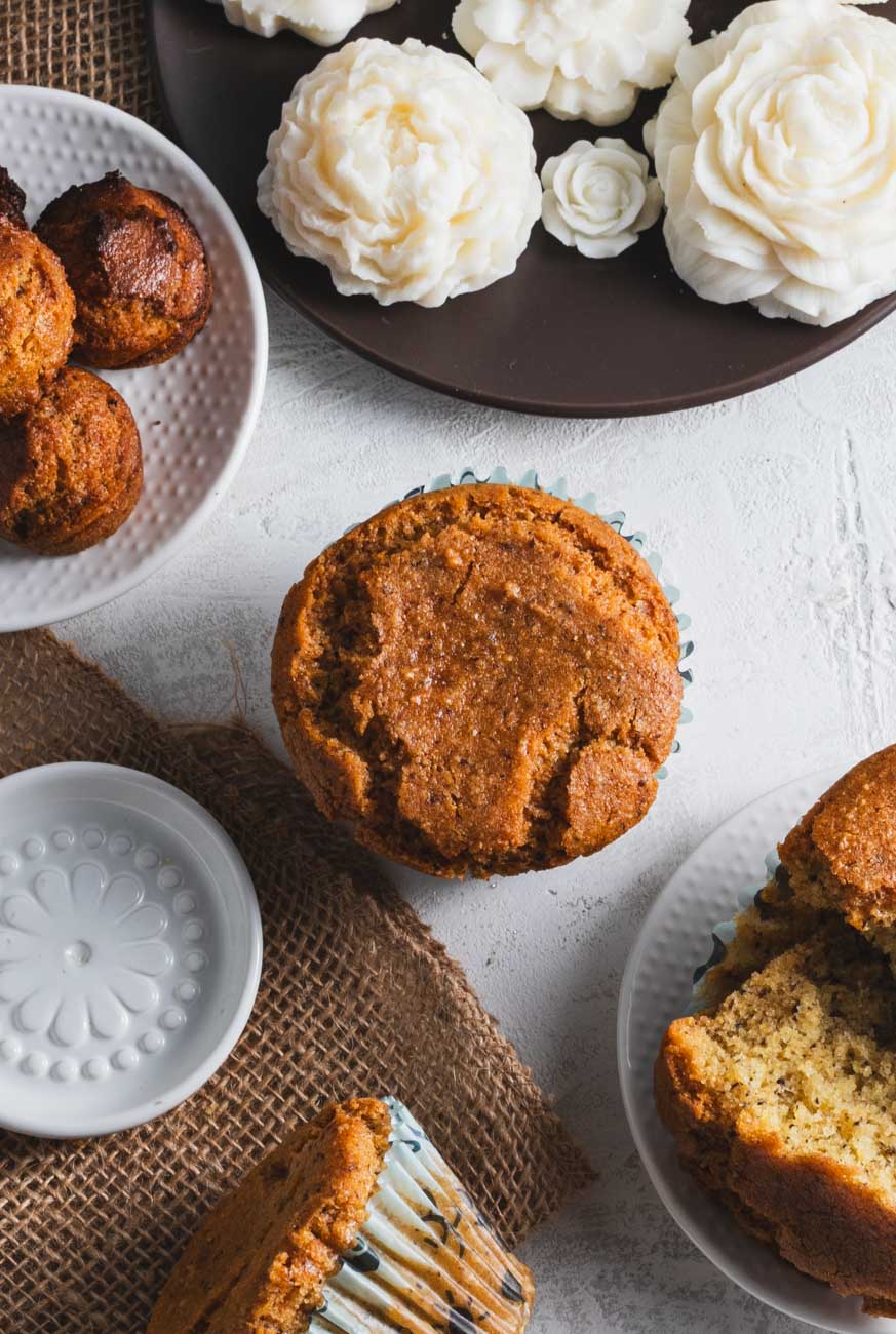 A top-down view of a large golden-brown corn muffin on a white plate, surrounded by mini corn muffins and white decorative flowers.