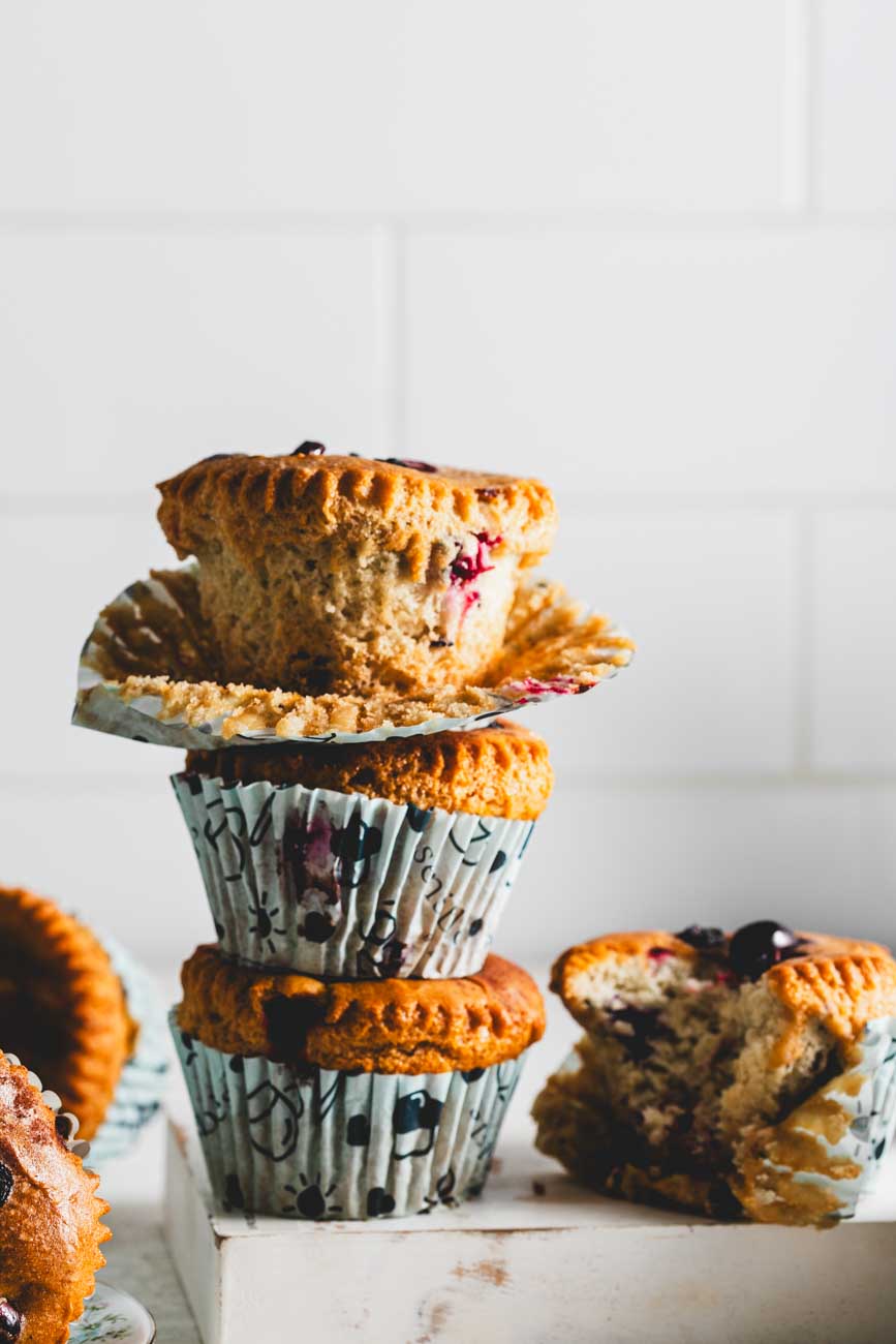 Stack of cinnamon pumpkin muffins with cranberries, showing one partially unwrapped muffin on top and a bitten muffin on the side, placed on a white surface.