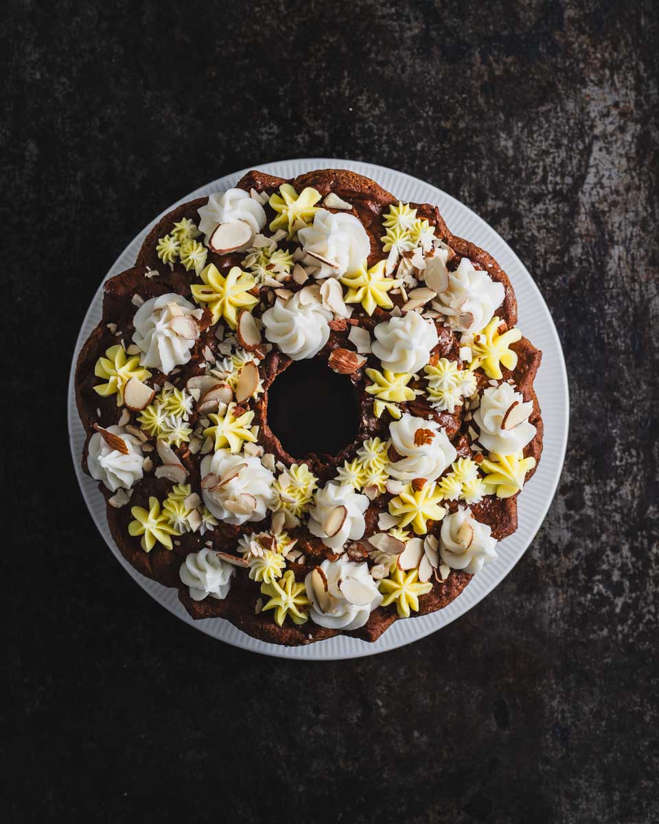Top view of a beautifully decorated lemon almond bundt cake on a white plate. The cake is adorned with swirls of white and yellow frosting and sprinkled with almond slices.