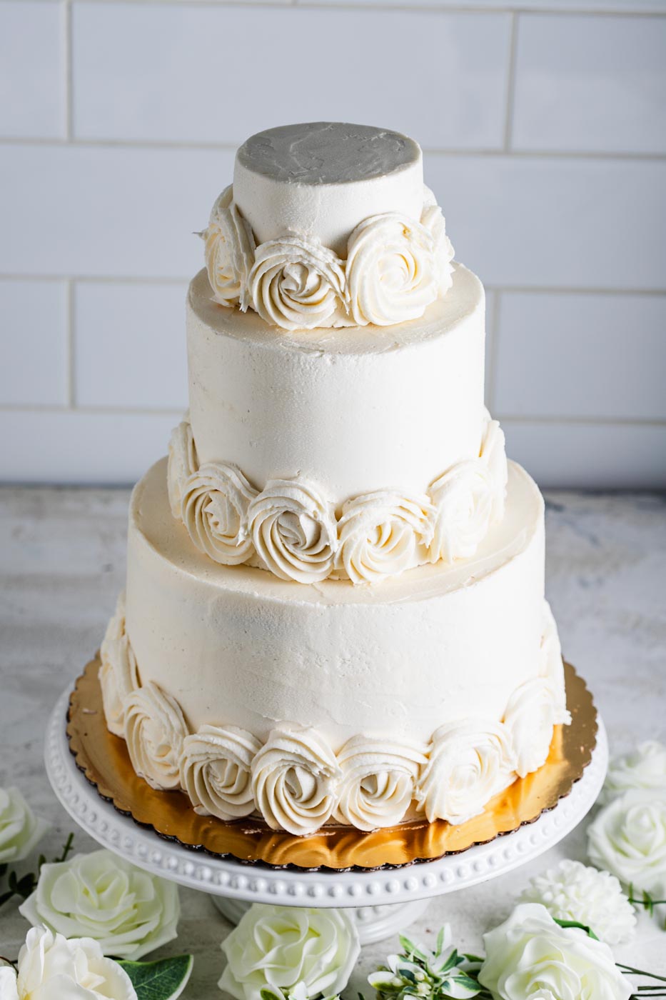 An elegant three tiered wedding cake with white frosting and white swirled frosting flowers around the base of each tier. There are decorative white flowers around the cake.