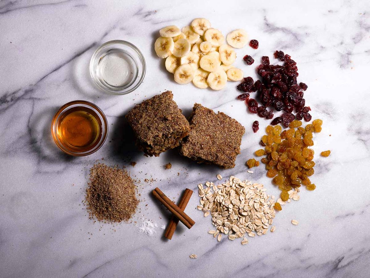 Ingredients for a breakfast protein bar arranged on a marble surface, including banana slices, dried cranberries, golden raisins, oats, cinnamon sticks, ground flaxseed, and maple syrup. Two protein bars are placed in the center.