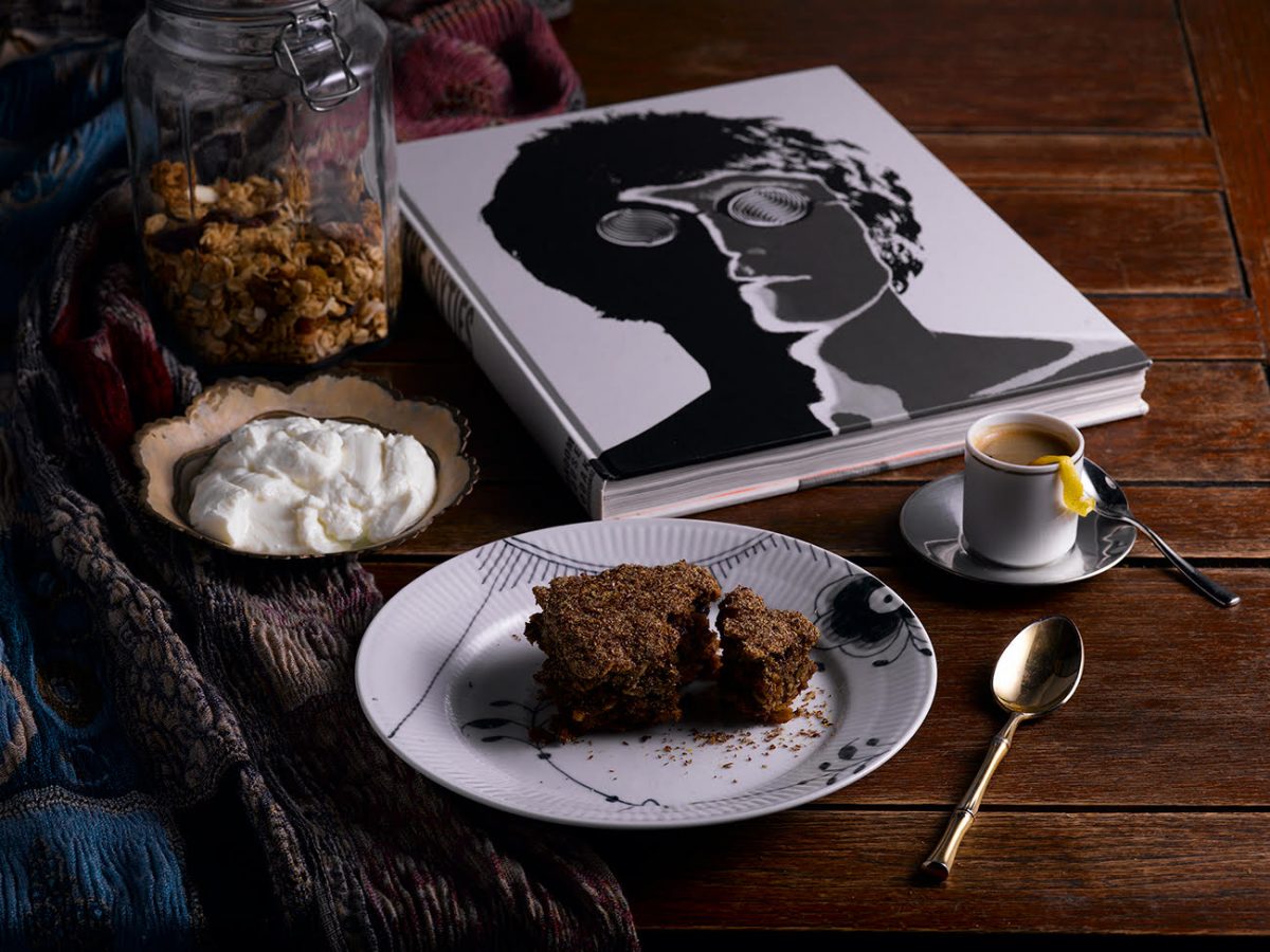 Breakfast protein bar served on a white plate with a cup of espresso and a dollop of whipped cream. The meal is accompanied by a jar of granola and a stylish black-and-white book on a wooden table.