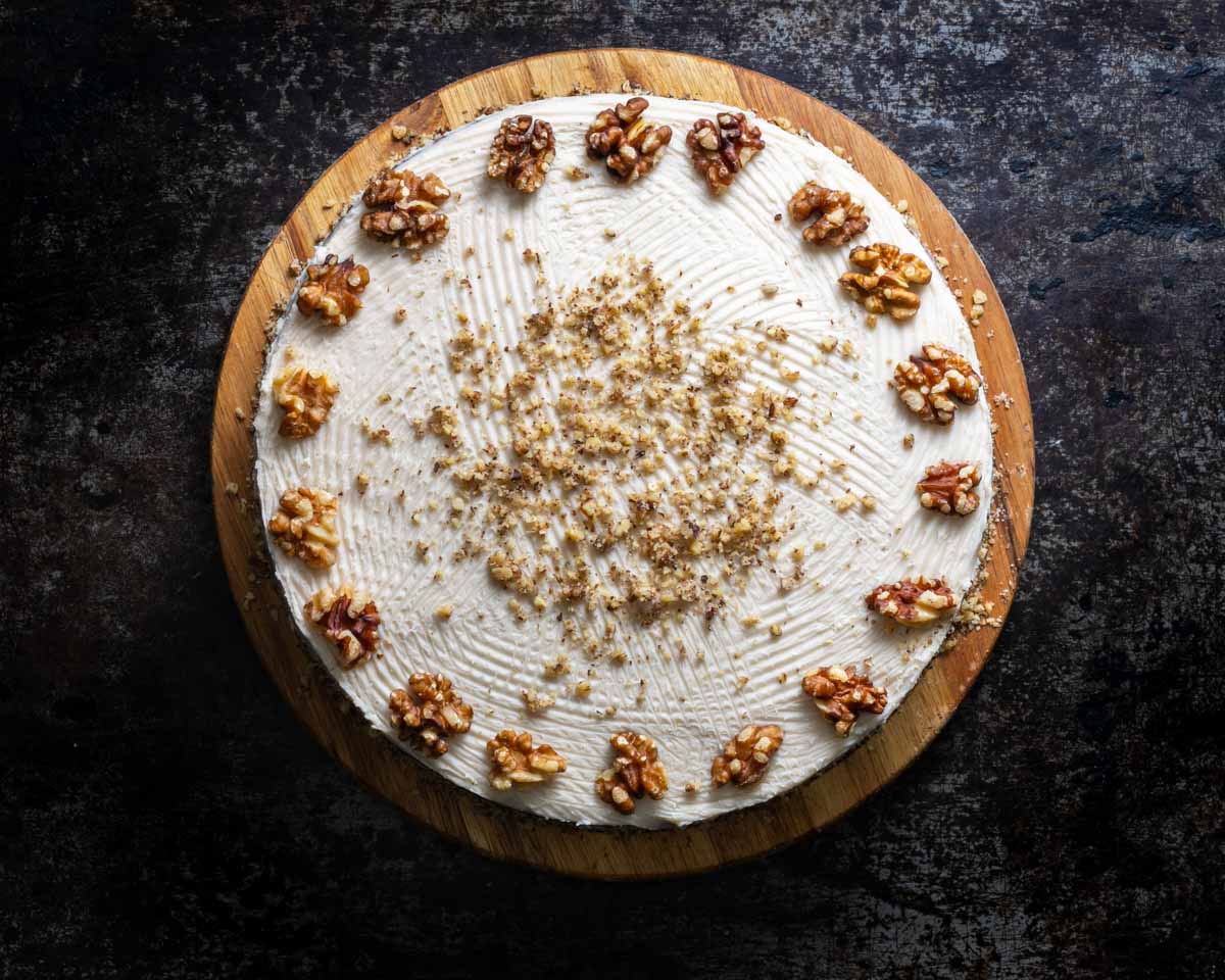 Top view of a frosted banana walnut cake topped with walnuts around the edges and finely chopped nuts in the center set on a wooden cake stand against a dark textured background.