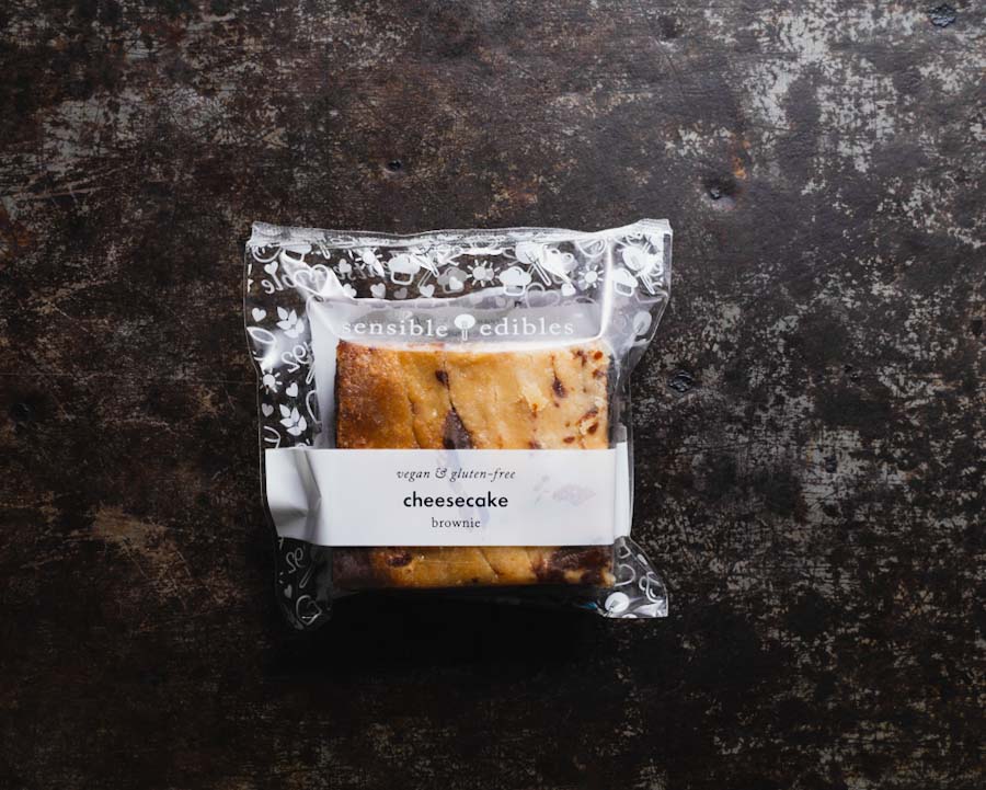 Packaged cheesecake brownie from Sensible Edibles, wrapped in clear packaging with a white label on a dark rustic surface.