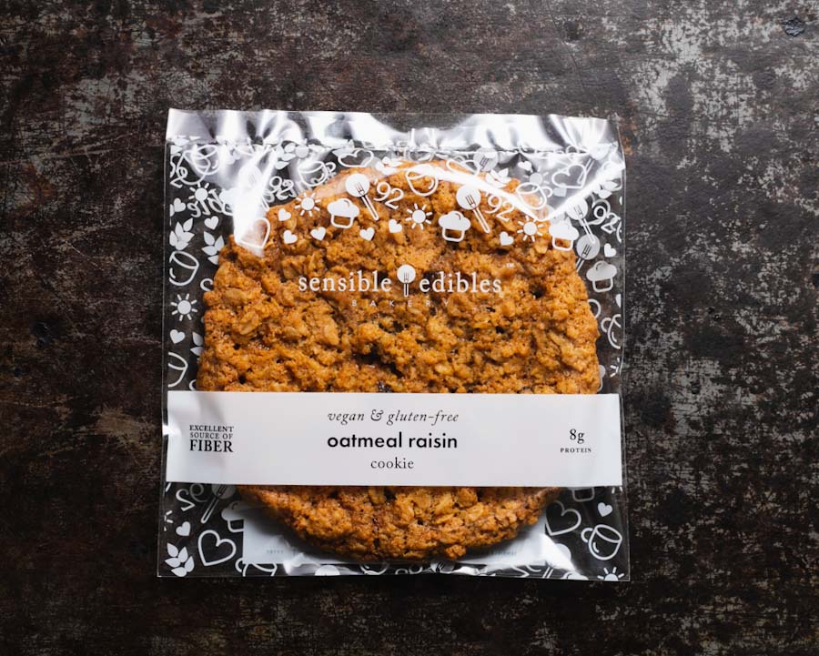 Packaged oatmeal raisin cookie from Sensible Edibles Bakery, labeled as vegan and gluten-free, on a dark background.