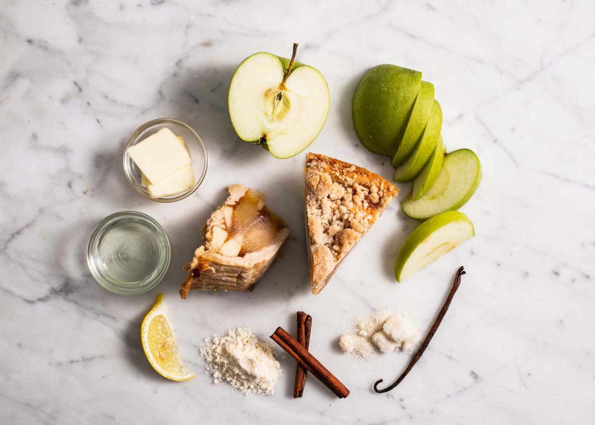 Two slices of mini apple pie on a marble surface with apple slices, lemon wedge, vanilla bean, and cinnamon sticks.