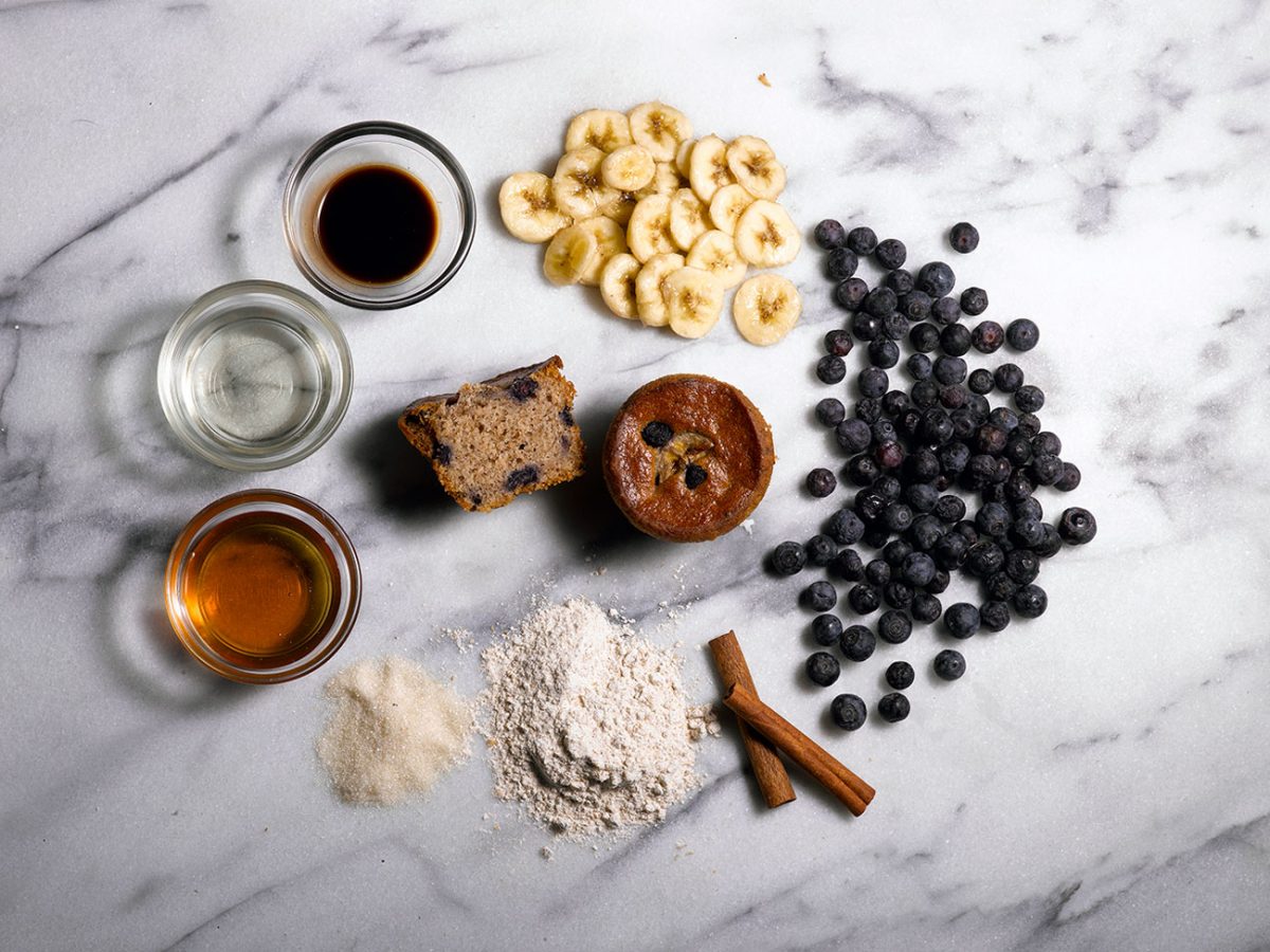 Banana blueberry muffin surrounded by blueberries, bananas, and cinnamon sticks on a white marble background.