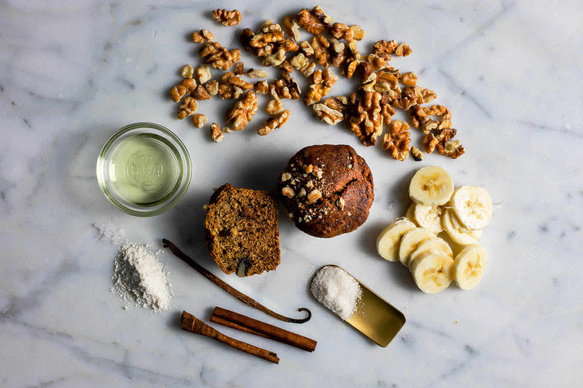Flat lay of ingredients for vegan and gluten-free banana walnut muffins, including a muffin in the center surrounded by banana slices, walnuts, flour, cinnamon sticks, vanilla beans, and a small bowl of oil arranged on a marble surface.