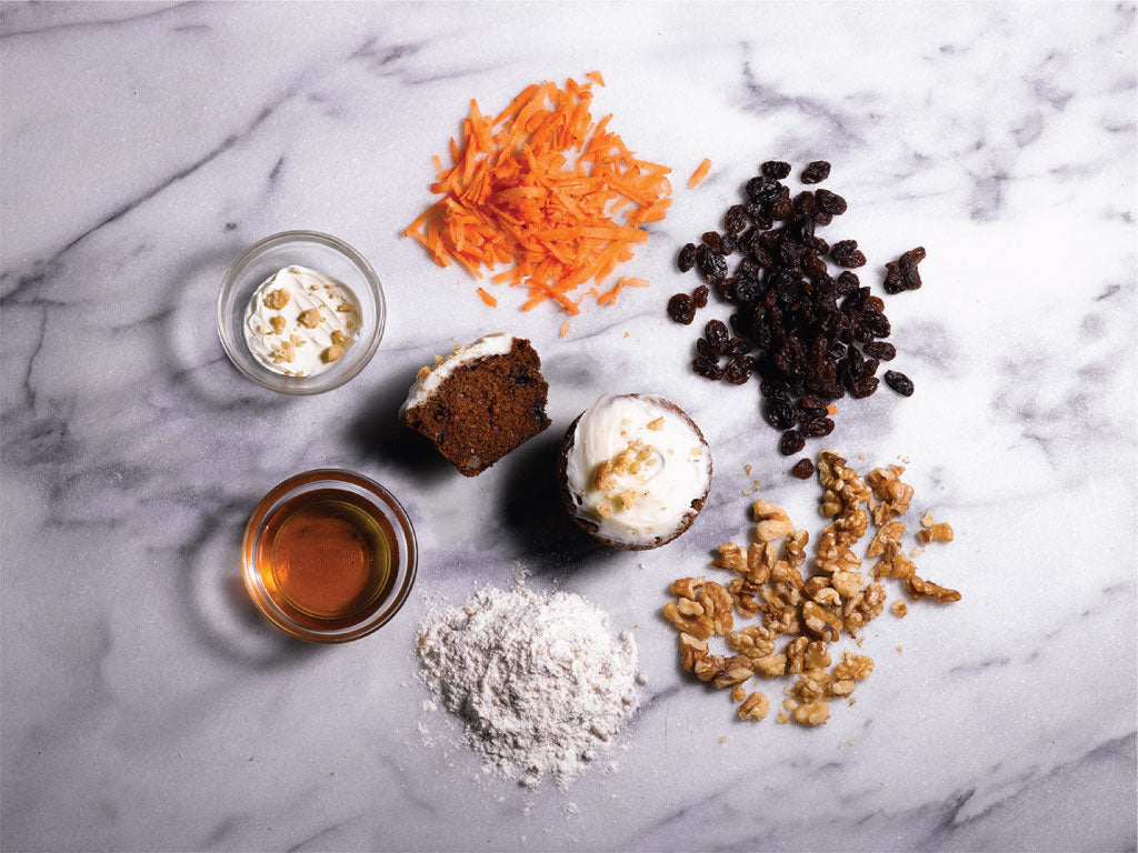 Ingredients for carrot walnut muffins, including grated carrots, raisins, walnuts, flour, maple syrup, and frosting, arranged around a sliced and a frosted muffin on a marble surface.