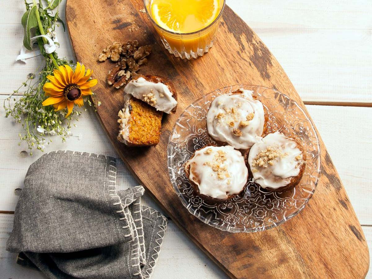Three carrot walnut muffins with creamy frosting and crushed walnuts on a glass plate, accompanied by a sliced muffin, a glass of orange juice, a yellow flower, and a grey napkin on a wooden board.