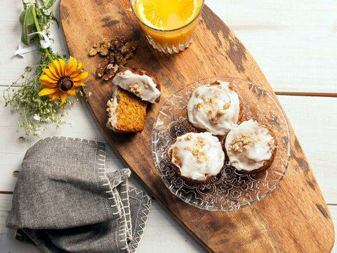 Three carrot walnut muffins with creamy frosting and crushed walnuts on a glass plate, accompanied by a sliced muffin, a glass of orange juice, a yellow flower, and a grey napkin on a wooden board.