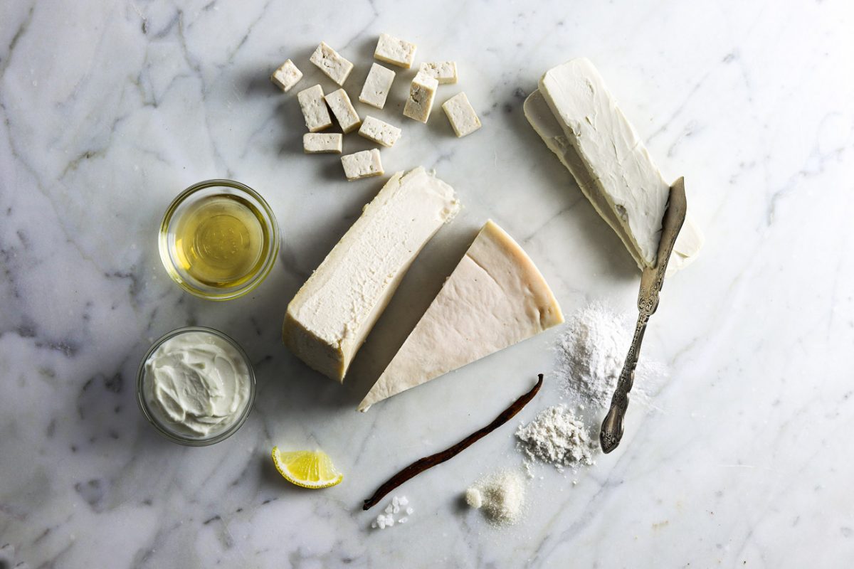 Ingredients for cheesecake, including tofu, vegan cream cheese, lemon, vanilla bean, flour, sugar, and vegetable oil, arranged around slices of cheesecake on a marble surface.