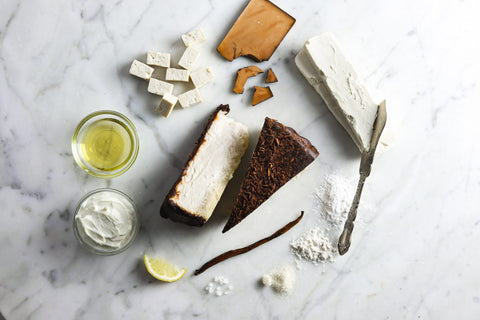 Ingredients for chocolate cheesecake, including tofu, vegan cream cheese, lemon, vanilla bean, flour, sugar, chocolate, and vegetable oil, arranged around a slice of cheesecake on a marble surface.