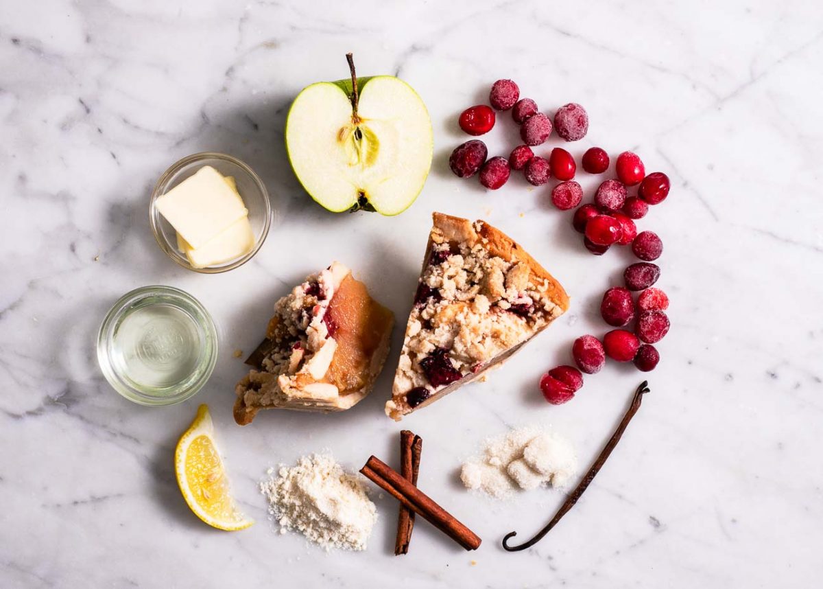 Two slices of cranberry apple pie with ingredients: apple slices, cranberries, butter, flour, cinnamon sticks, and vanilla bean on a marble surface.