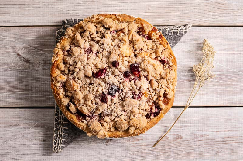 Whole cranberry apple pie with a crumb topping on a wooden surface, served on a gray cloth.