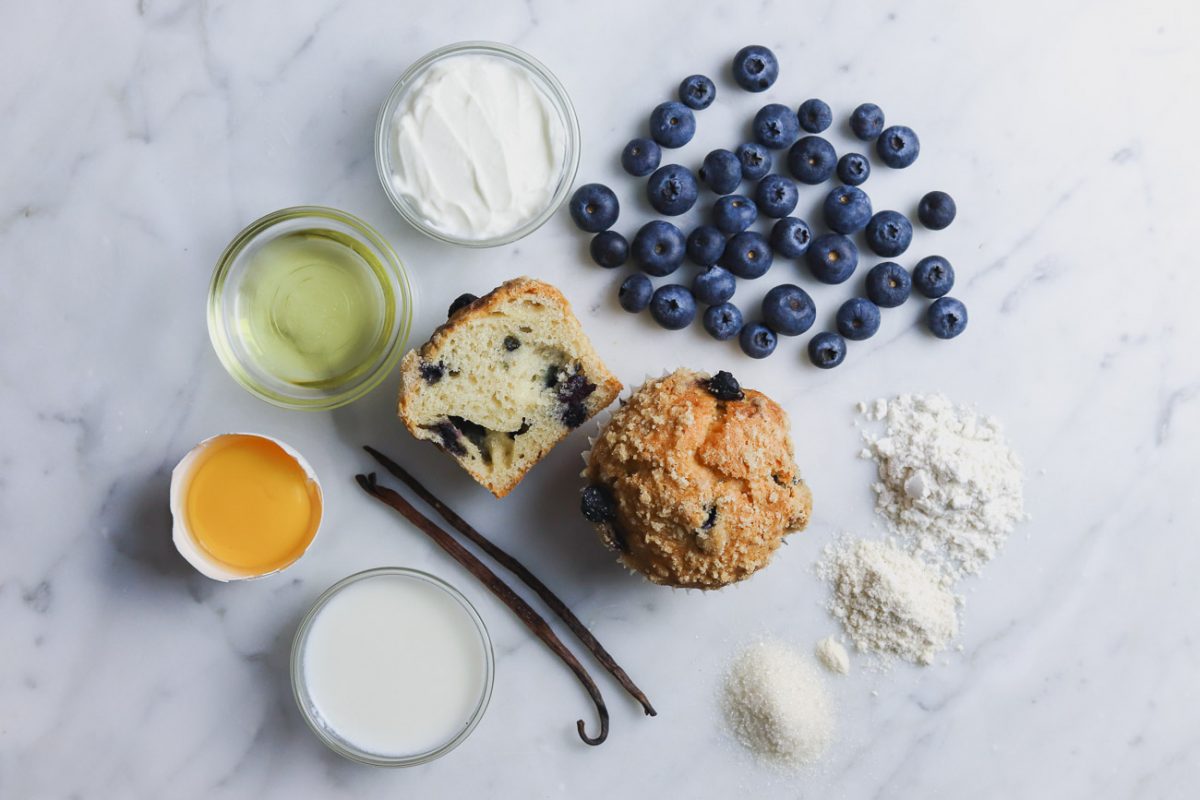 Flat lay of ingredients, including a blueberry crumb muffin in the center surrounded by blueberries, flour, sugar, vanilla beans, milk, egg, oil, and yogurt arranged on a marble surface.