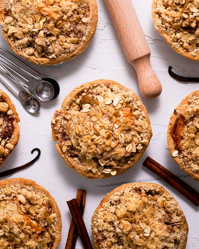 Mini apple crumb pies on a white surface with cinnamon sticks and vanilla beans