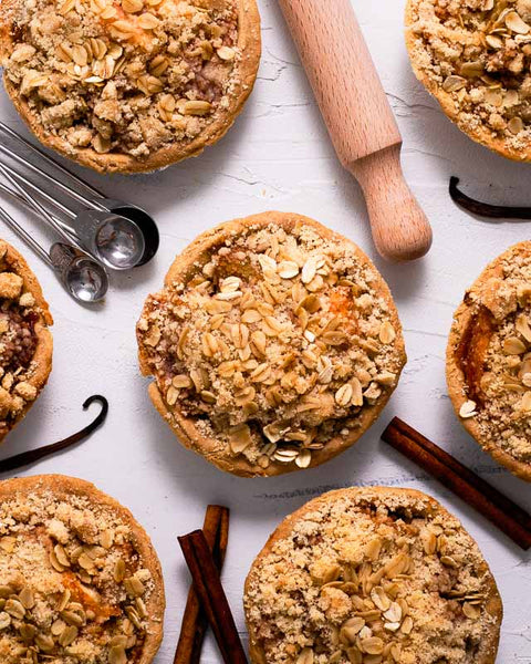 Mini apple crumb pies on a white surface with cinnamon sticks and vanilla beans