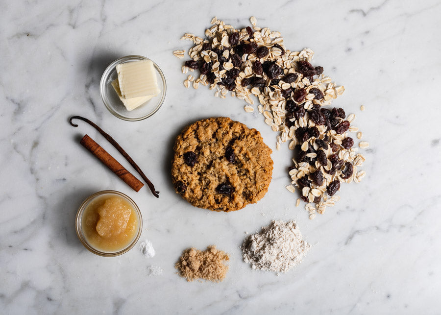 Oatmeal raisin cookie surrounded by ingredients including oats, raisins, butter, vanilla bean, cinnamon sticks, applesauce, brown sugar, and flour on a marble surface.