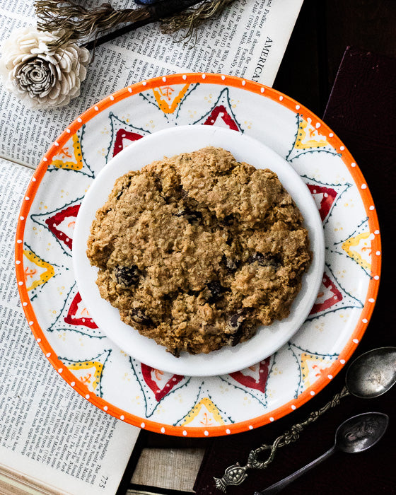 A single oatmeal raisin cookie on a colorful patterned plate, featuring vintage papers and spoons.