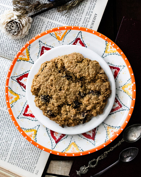 A single oatmeal raisin cookie on a colorful patterned plate, featuring vintage papers and spoons.