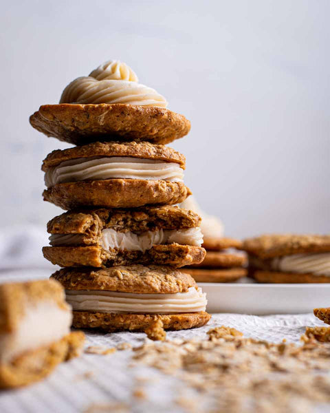 A stack of oatmeal cream whoopie pies with creamy filling, highlighting the soft texture and oatmeal exterior, with more whoopies in the background on a white surface.