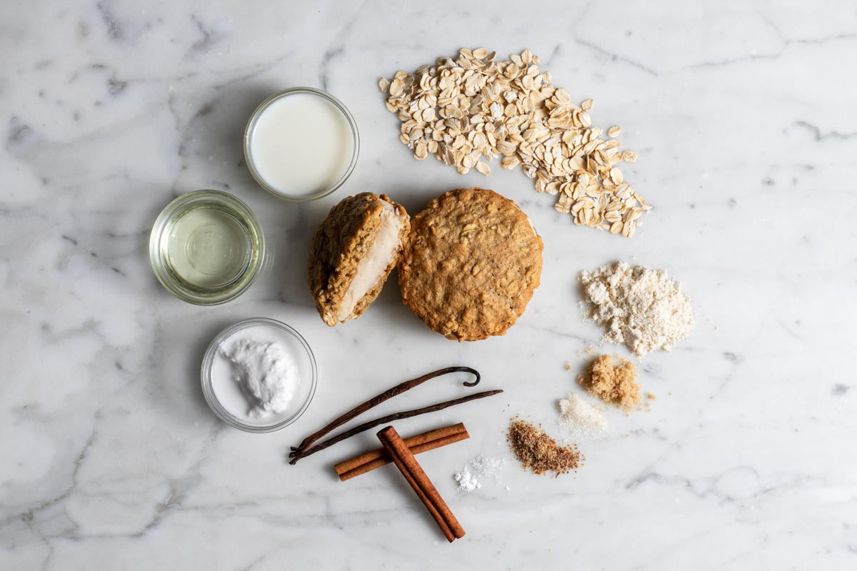  Two oatmeal cream whoopie pies surrounded by ingredients including oats, milk, oil, coconut oil, flour, cinnamon sticks, and vanilla bean on a marble surface.