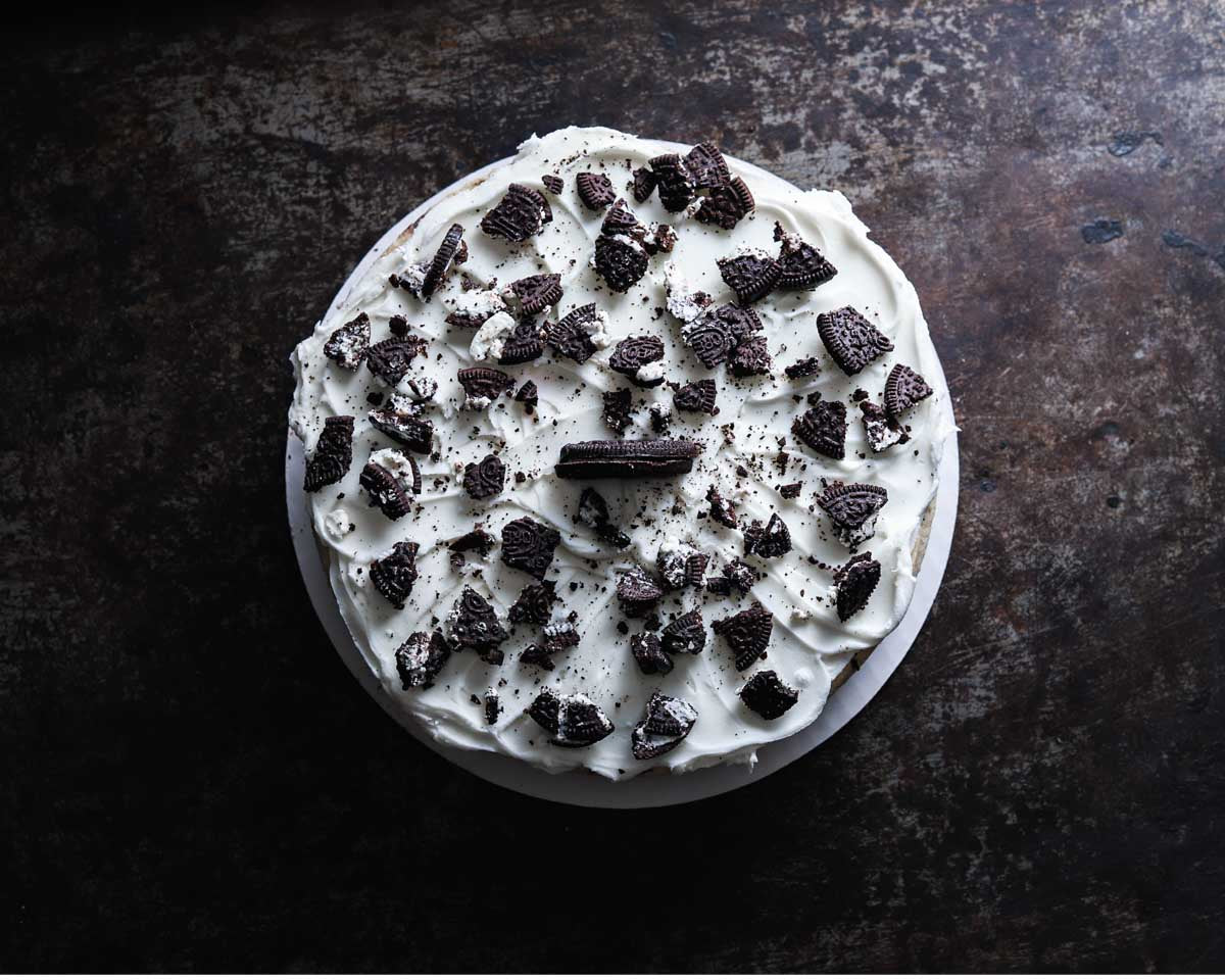 Top-down view of an Oreo cheesecake, covered with whipped cream and generously sprinkled with crushed Oreo cookies on a dark, textured background.