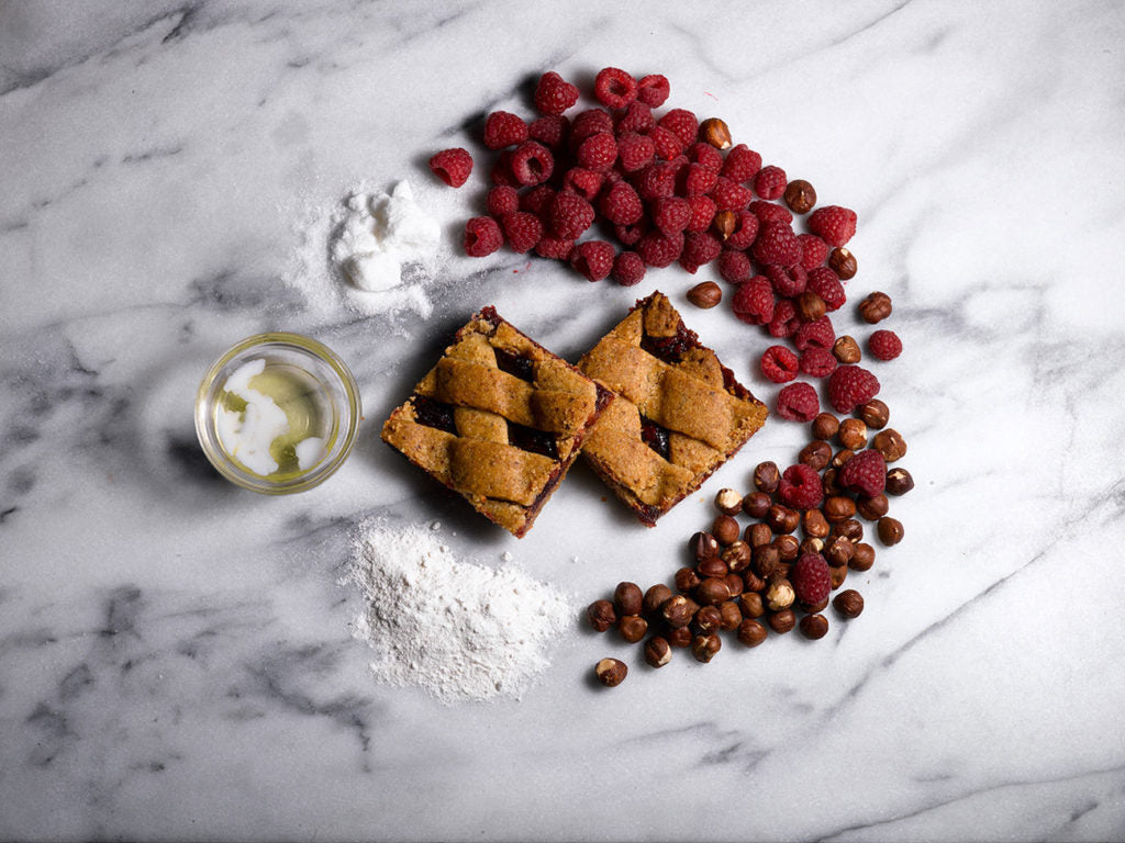Two raspberry hazelnut bars on a marble surface, surrounded by fresh raspberries, hazelnuts, and flour.